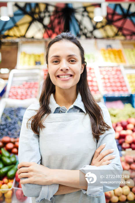 Positive confident young female grocer in apron crossing arms on chest and looking at camera while standing against fresh food stall at farmers market