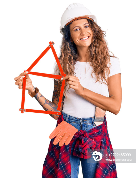 Young hispanic woman with tattoo wearing architect hardhat holding build project looking positive and happy standing and smiling with a confident smile showing teeth