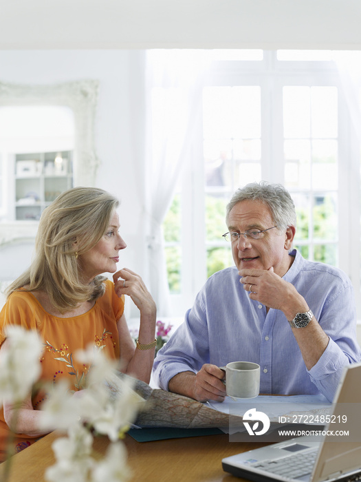 Mature couple with map and laptop sitting at dining table in home