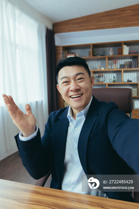 Cheerful and funny Asian businessman looks at the camera of a smartphone, talks on a video call, a man works in the office, waves a congratulatory gesture