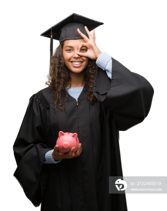 Young hispanic woman wearing graduation uniform holding piggy bank with happy face smiling doing ok sign with hand on eye looking through fingers