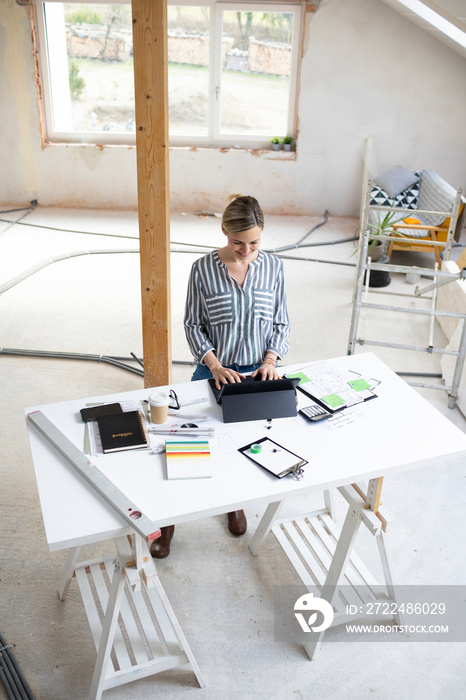 pretty young architect working on construction site in a loft and standing by a high table wearing glasses and grey and white striped top with coffee and notebook