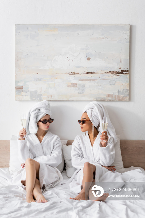 full length of barefoot interracial women in bathrobes and trendy sunglasses holding champagne and looking at each other on bed.
