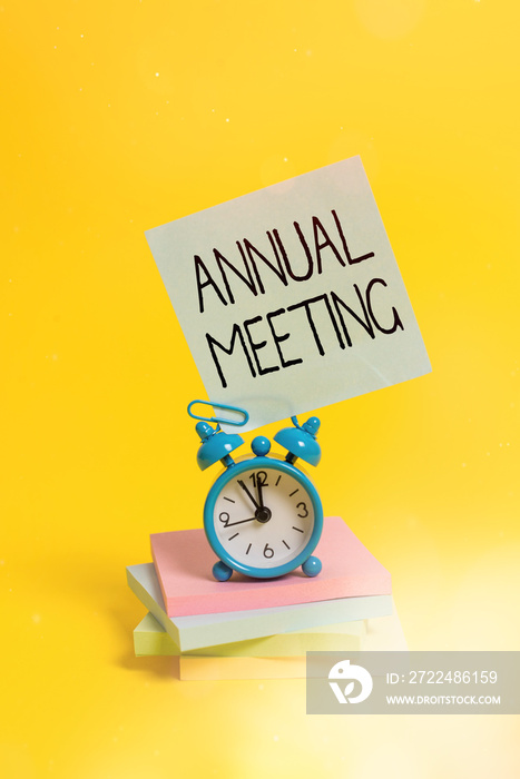 Handwriting text Annual Meeting. Conceptual photo yearly meeting of the general membership of an organization Metal alarm clock blank sticky note stacked notepads colored background