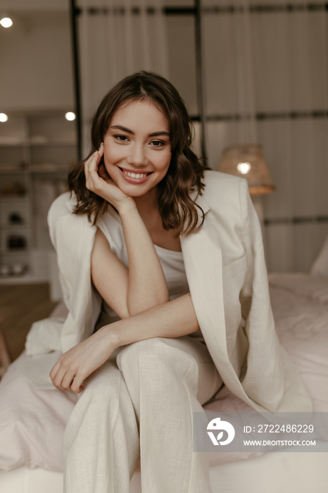 Brown-eyed woman in beige pants and linen jacket smiles widely and looks into camera. Brunette curly lady in suit sits on soft bed.