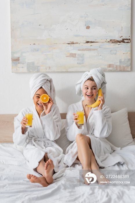 full length of cheerful interracial women with fruits and orange juice having fun while sitting on bed.