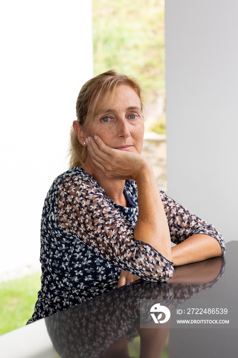 Portrait of a middle-aged woman with blond hair in a ponytail with a window with a green lawn and a white wall behind her