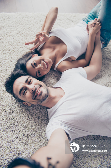 Cheerful playful ltino sister and brother are taking selfie and making funny grimaces, gesturing. They lie on the floor on beige carpet in white casual outfits indoors at home