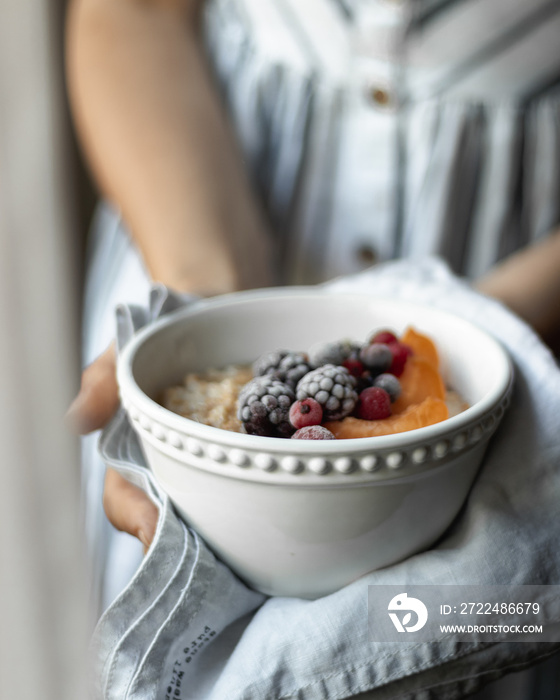 Girl hands holding oatmeal porridge with frozen berries, almonds and caramelized apricots in white ceramic bowl. Healthy breakfast. Clean eating, detox diet. Vegetarian, raw, vegan concept