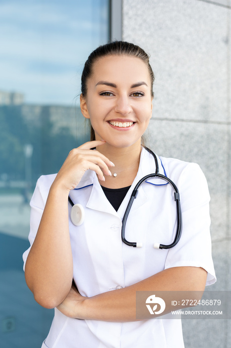 Healthcare, medicine, profession and people concept - portrait of a happy European white-haired doctor girl with her hand at her face in a medical gown, wearing a stethoscope outside in sunny weather