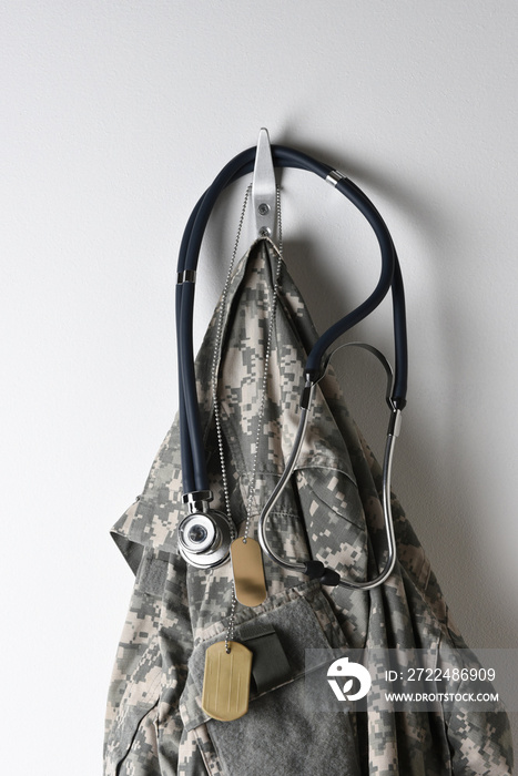 A set of military dog tags, Stethoscope and camouflage field jacket hanging from a hook on a blank wall.