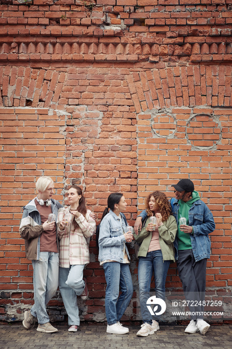 Group of multicultural teenage guys and girls in casualwear standing against brick wall, having soda and chatting after college