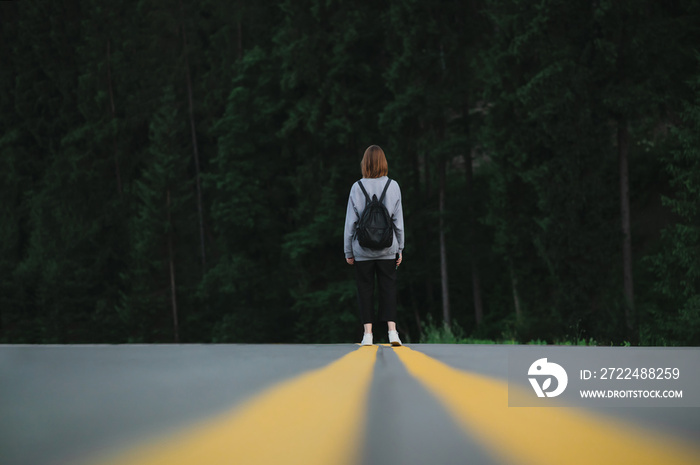 Back view on a female hiker walking down the asphalt road in the woodland. Tourist woman with a backpack standing in the middle of the highway near the fir forest. Travel concept.