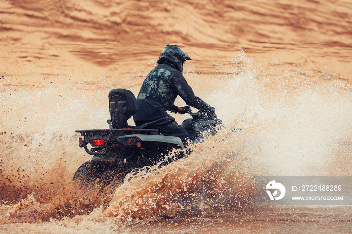 Quad rider go fast at the sands dunes through water. Quad Bike driver ride at the desert with a splash of water