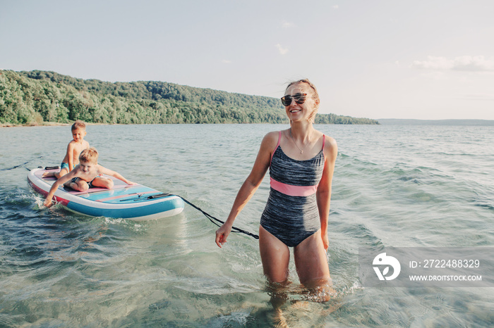 Caucasian woman parent riding kids children boys on paddle sup surfboard in water. Modern outdoor summer fun family activity. Individual aquatic recreation sport hobby. Healthy lifestyle.