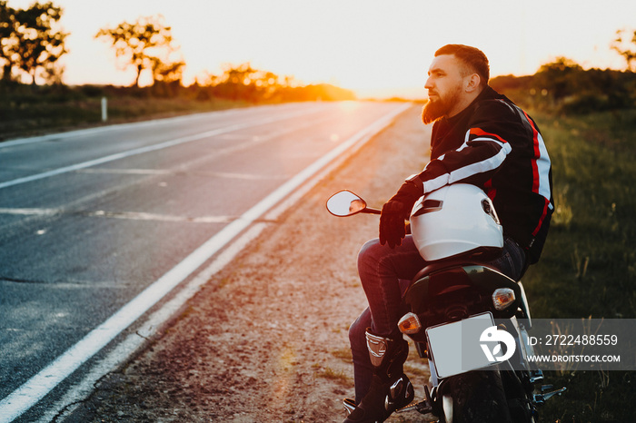 Side view portrait of a confident adult young caucasian male biker sitting on his motorcycle and looking away with a hand on his helmet near the road while traveling against sunset.