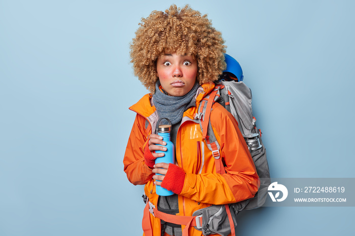 Worried curly female explorer has frozen face holds thermos with hot beverage carries heavy rucksack wears orange windbreaker and scarf discovers something new isolated over blue background.