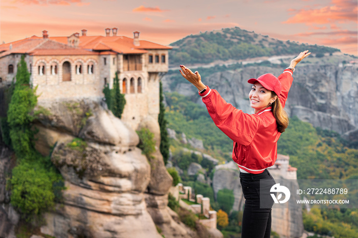 Happy traveler girl with raised arms standing on top of a cliff and admires view of the famous Meteora monasteries. Discover wonders of the world and tourist experience