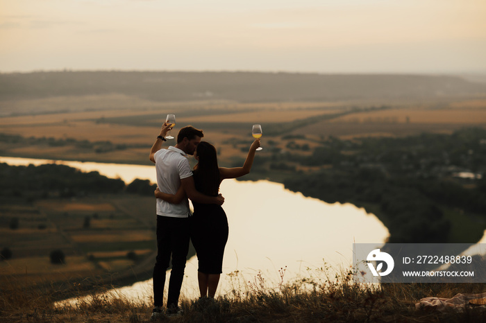 Romantic couple kissing on a mountain and holding glasses of wine at evening. Happy gorgeous elegant couple kissing on summer picnic.