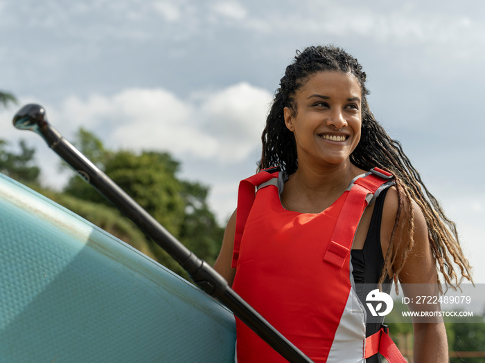 Portrait of smiling woman carrying paddleboard and oar