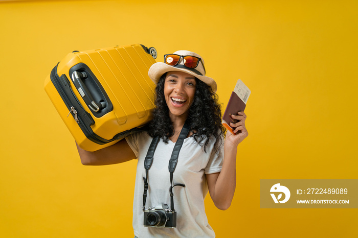 Happy Traveler curly Latin woman wears white t-shirt hold suitcase bag, passport and boarding tickets isolated on yellow background.