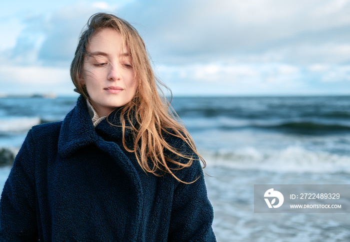 Portrait of Young beautiful woman braving a cold winter day at the seaside. Happy female with long hair in turquoise coat resting during long solo trip standing on the beach and enjoying the sea wave.