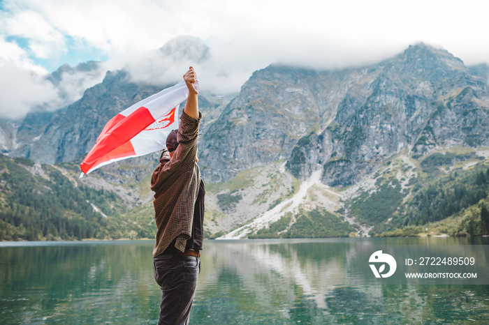 man covered in poland flag standing on the edge looking at lake in mountains