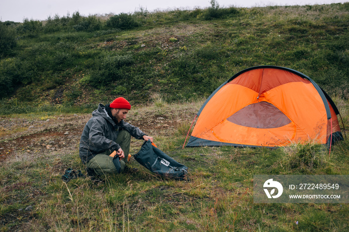 Young male explorer tourist unpacks his dry bag or backpack with camping gear, sleeping matress and hiking food, sets up camp next to tent in wild national park forest or field. Adventure lifestyle