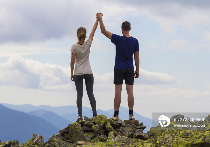 Back view of young tourist couple, athletic man and slim girl standing with raised arms holding hands on rocky mountain top enjoying fantastic panorama. Tourism, traveling and climbing concept.