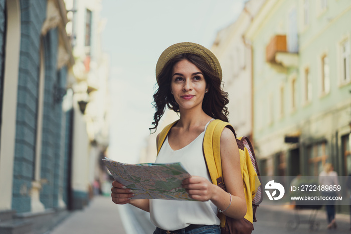 The brunette girl-traveler with a map of the city and a backpack in the street. With a cute smile. And an architecture on the background.