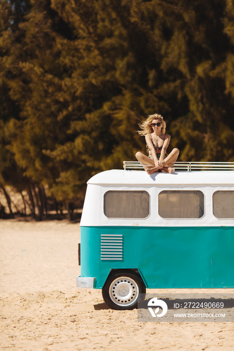 Happy young lady with long curly hair sitting on blue and white mini bus with crossed legs during summer days. Charming woman in sunglasses and swimwear relaxing on beach of Hainan island.