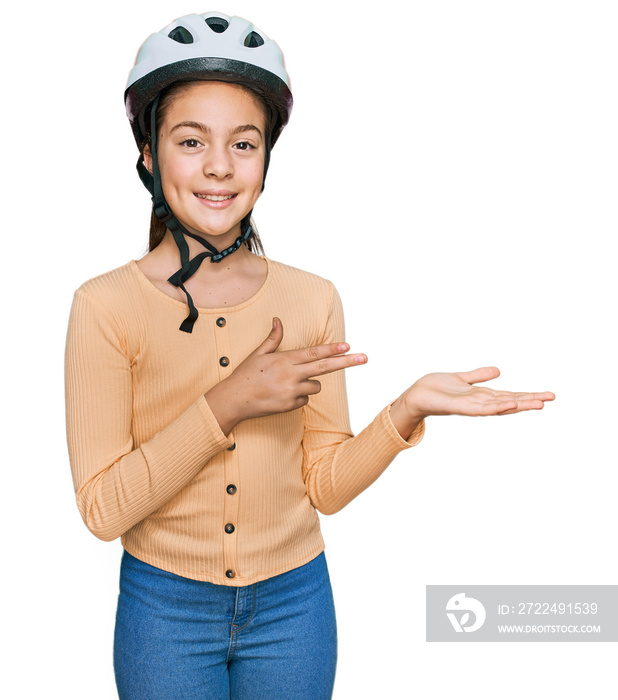 Beautiful brunette little girl wearing bike helmet amazed and smiling to the camera while presenting with hand and pointing with finger.