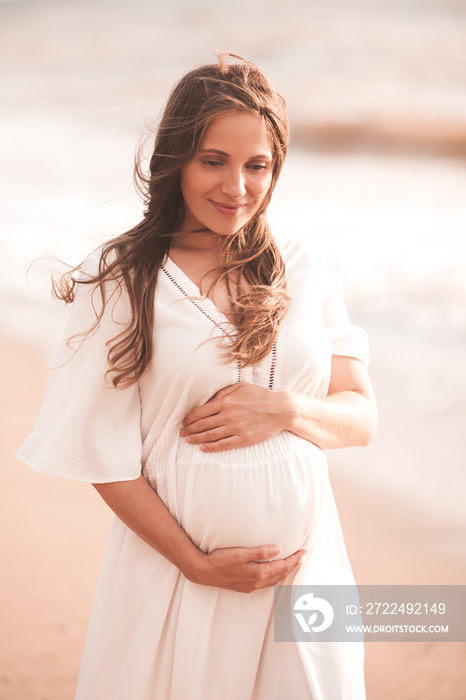 Blonde pregnant woman 24-26 year old wearing white dress holding tummy walking at beach outdoors. Motherhood. Maternity. Healthy lifestyle. Happiness.