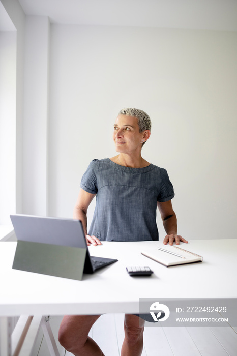 middle-aged businesswoman standing by white high table and working on tablet and wearing grey-blue dress and with short light hair