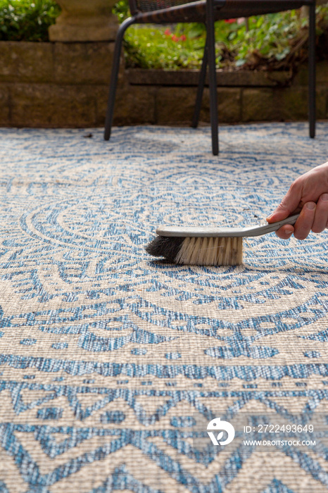 Man brushing an outdoor rug in a garden with a hand brush.  Grey and blue design