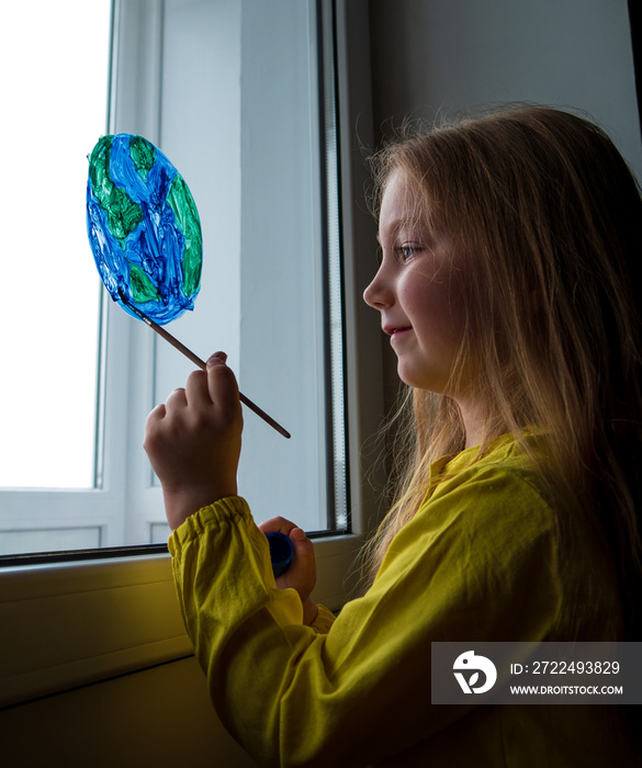 Cute little girl painting planet on window at home. Happy Earth Day April 22 greeting message. Creative family leisure lockdown new reality. Ecology Saving environment conscious consumption concept.