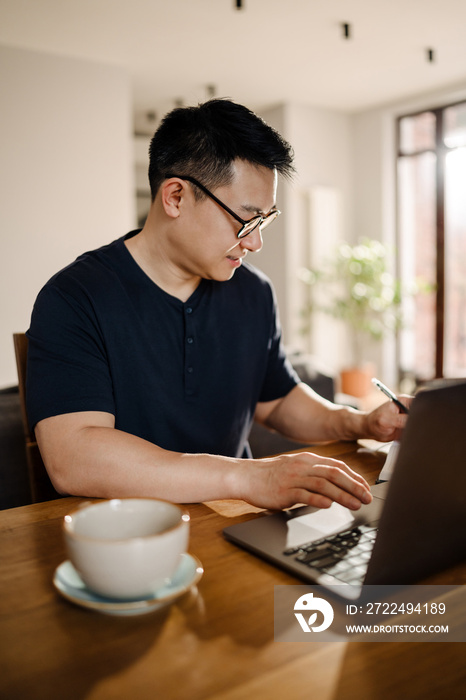 Brunette adult asian man smiling and using laptop computer at home