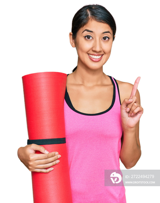 Beautiful asian young sport woman holding yoga mat surprised with an idea or question pointing finger with happy face, number one