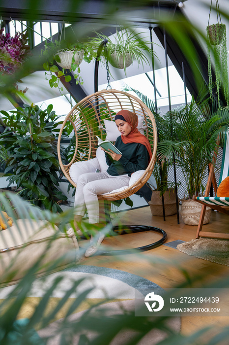 Woman wearing headscarf sitting in swing chair and reading book