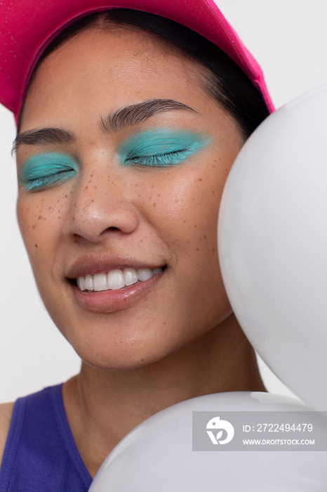 Woman with closed eyes, wearing blue makeup and sun visor next to white balloons