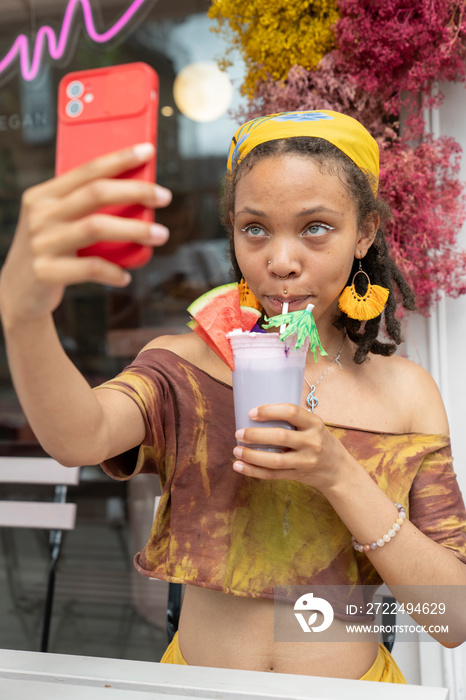 Young woman taking selfie while drinking smoothie in front of sidewalk cafe