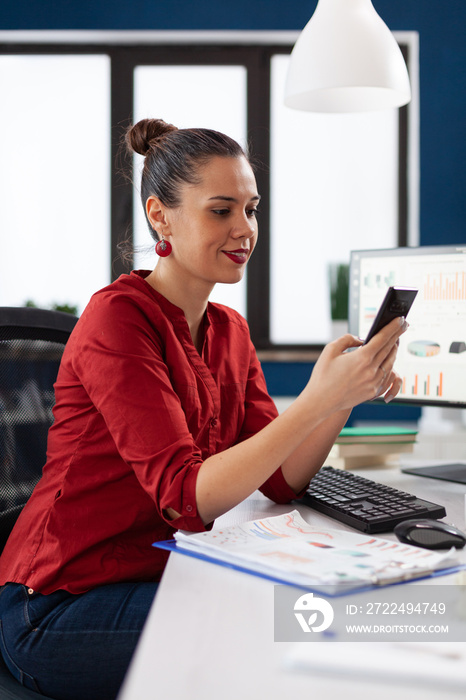 Manager using smartphone in startup business office. Businesswoman in red shirt texting sitting at desk. Company employee browsing internet on the phone in front of desktop computer.