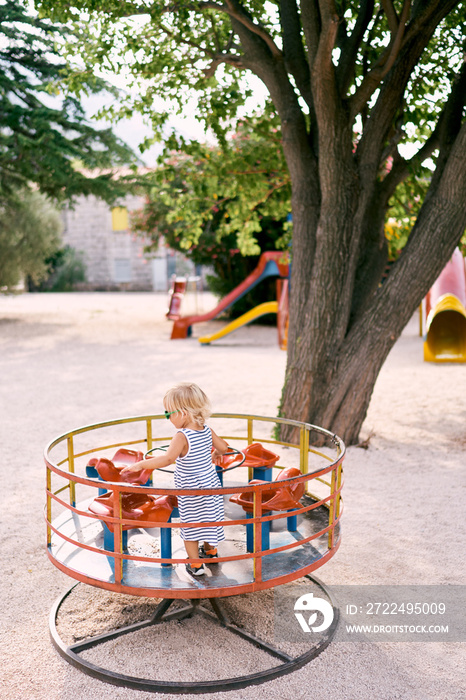 Little girl in sunglasses stands on a turntable swing. Back view