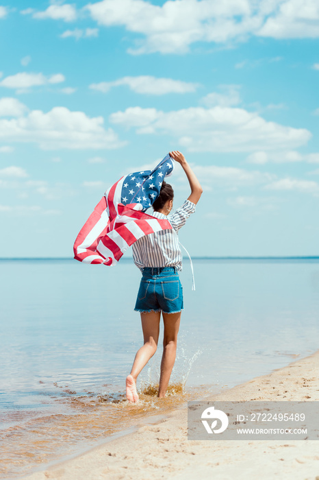 rear view of young woman running in sea water with american flag, independence day concept