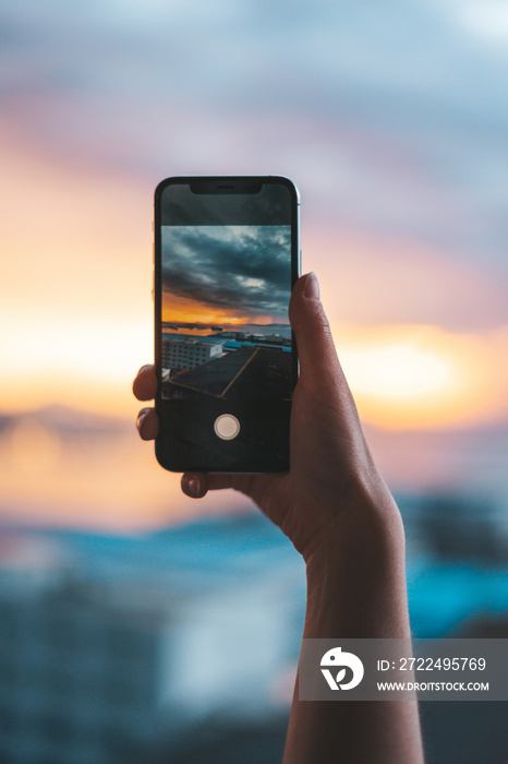 Hand holding a smartphone taking a photograph of the sea at sunset