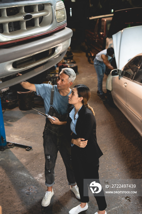 Senior male mechanic in uniform checking and diagnostic car while talking to young woman in business clothing with folding arms and informing about vehicle issue in garage while working