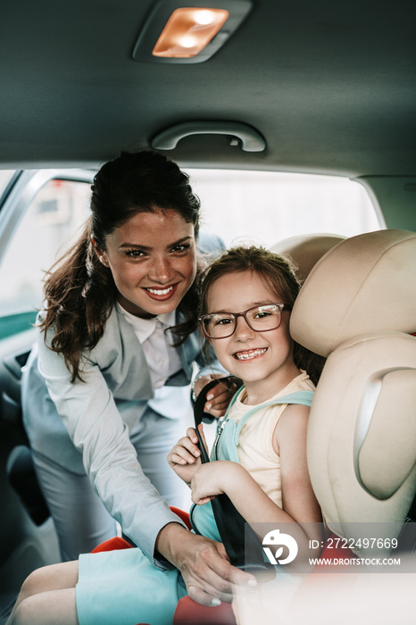 Young businesswoman helping her daughter to fasten seatbelts in the car while girl is sitting on a safety child car seat.