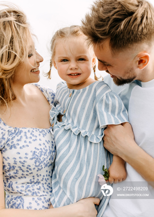 Happy young family close-up portrait outdoors. Mother and father hugging little daughter, smiling.