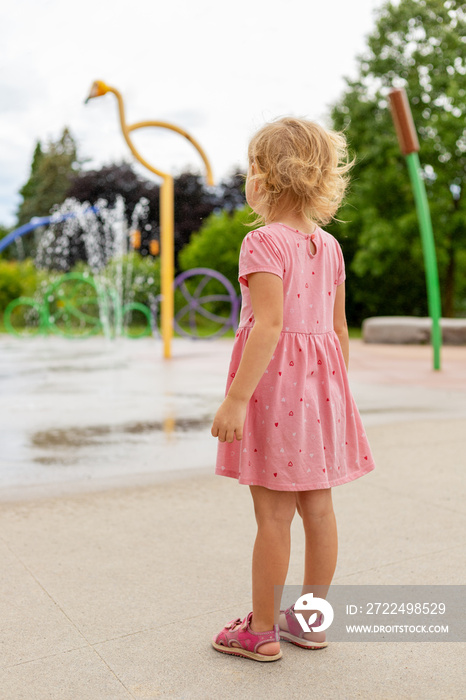 Little girl playing at splash pad playground in park in summer. Child from behind near fountain