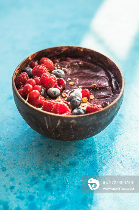 Frozen acai smoothie in coconut shell with raspberries, banana, blueberries,  fruit and granola on concrete background. Breakfast, healthy meal for summer vibes, top view, space for text